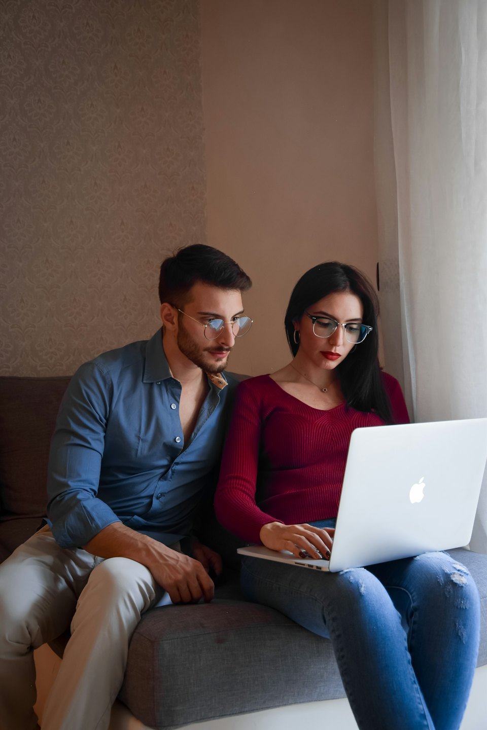 Para w okularach korzysta z srebrnego laptopa na szarej kanapie.